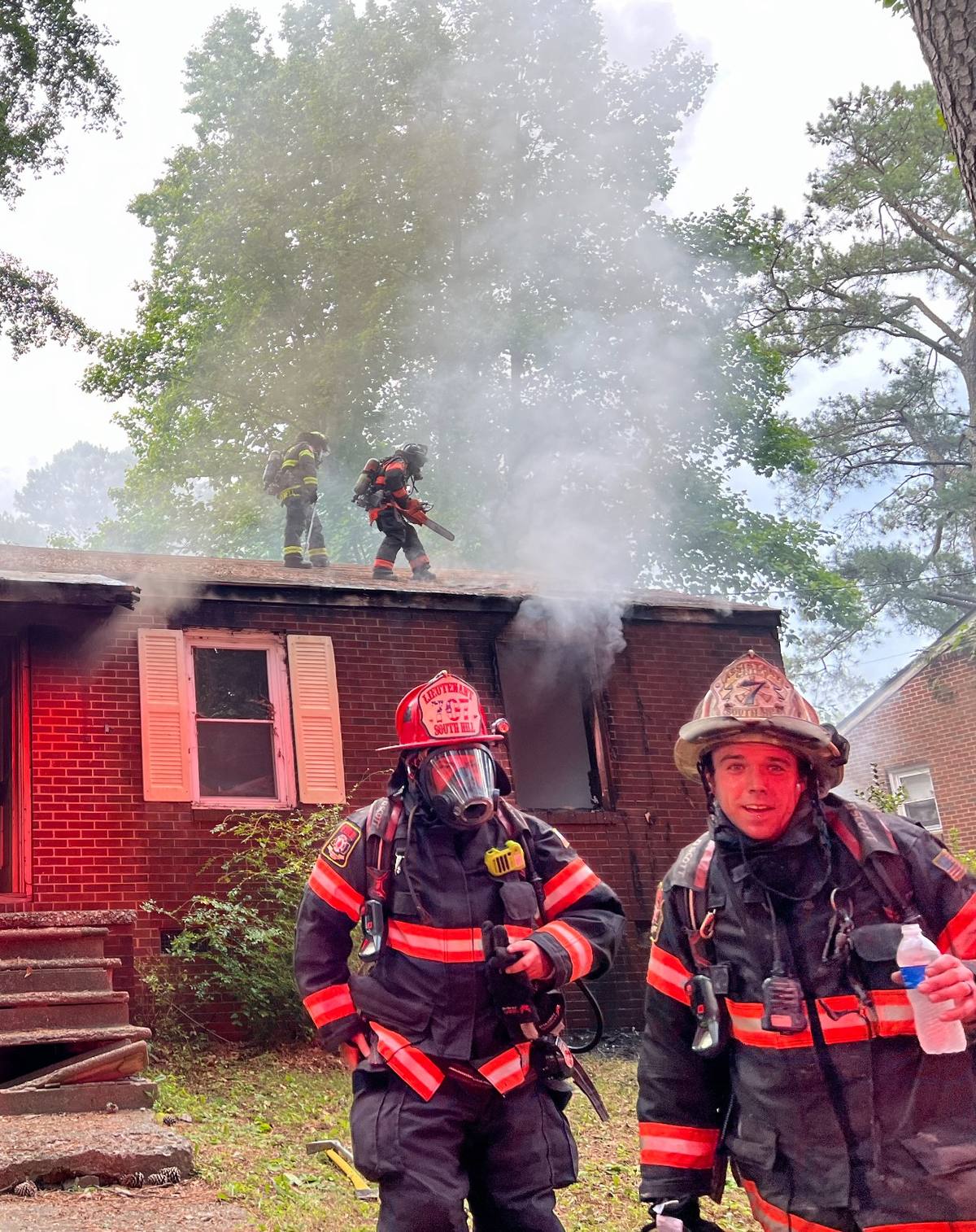 Two firefighters on air outside a working house fire while a crew member works the roof